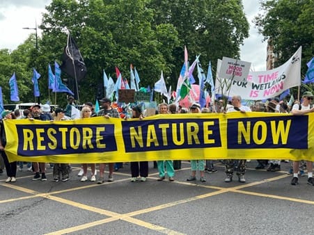 Photo of people marching on a road with a banner saying 'Restore Nature Now'